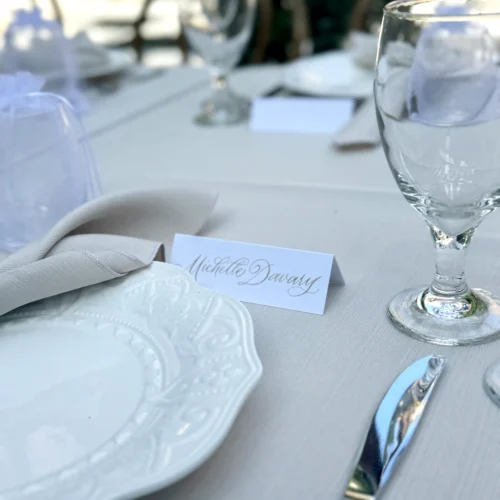 Elegant wedding place card featuring gold calligraphy by KeeKee Creates, displayed on a beautifully set table with glassware and fine tableware.