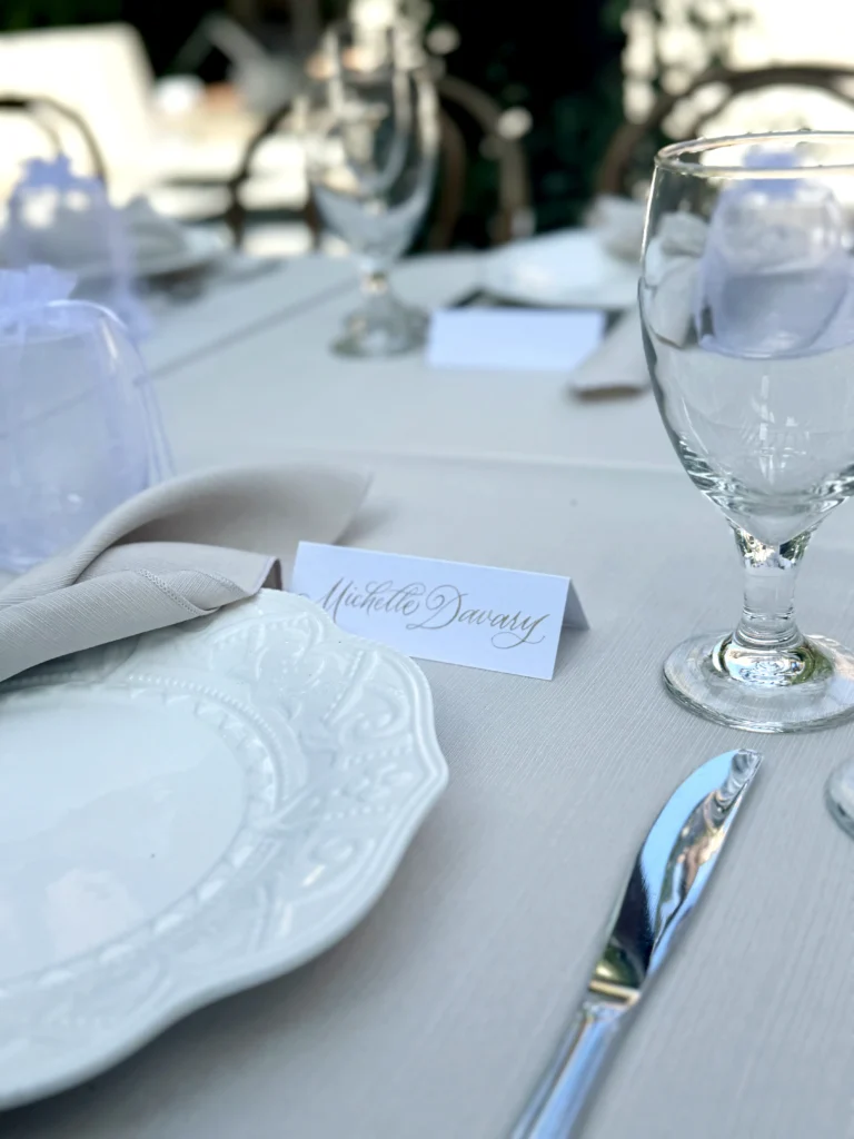 Elegant wedding place card featuring gold calligraphy by KeeKee Creates, displayed on a beautifully set table with glassware and fine tableware.