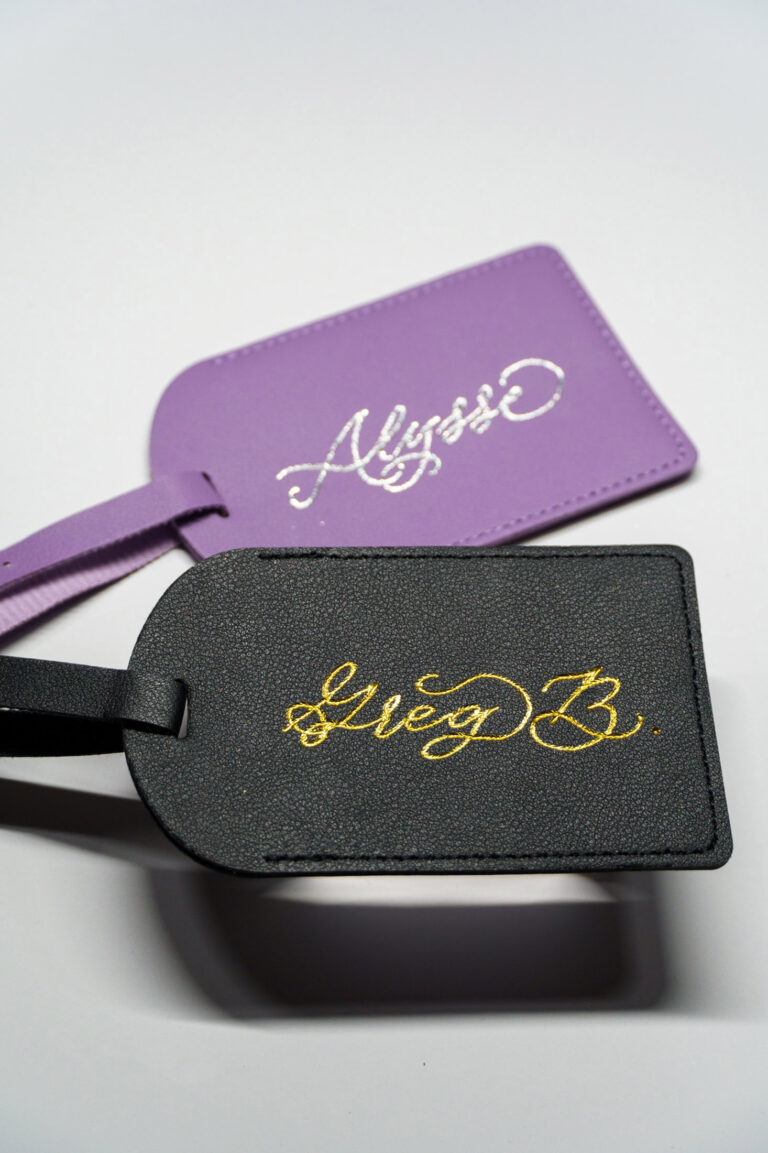 Custom leather luggage tags by KeeKee Creates featuring gold foil calligraphy names 'Alysse' and 'Greg B.' on purple and black tags