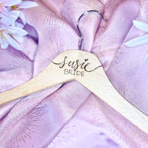 Personalized wooden wedding hanger with 'Susie Bride' calligraphy by KeeKee Creates, displayed on lavender fabric with white flowers